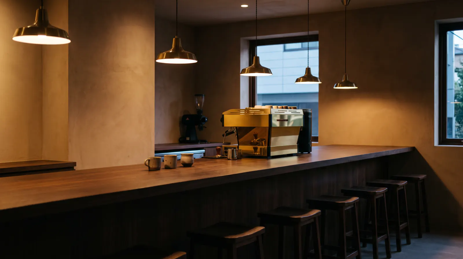 The café in the evening: a long dark wood counter, a brass espresso machine and warm pendant lights